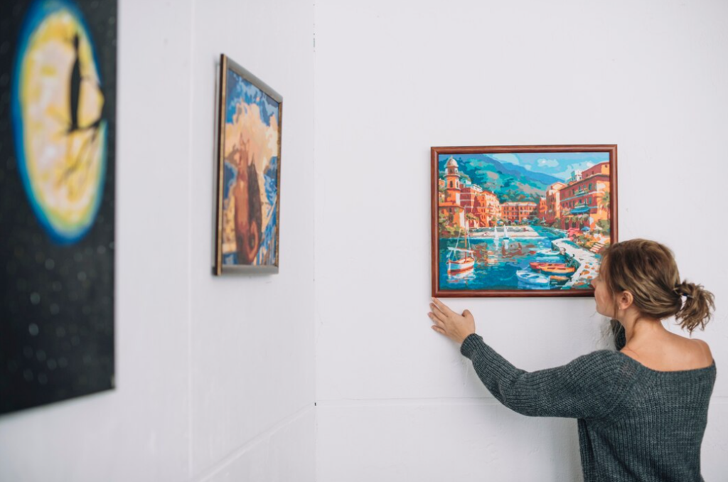 A woman in a grey sweater adjusts a vibrant painting of a coastal town on a white gallery wall