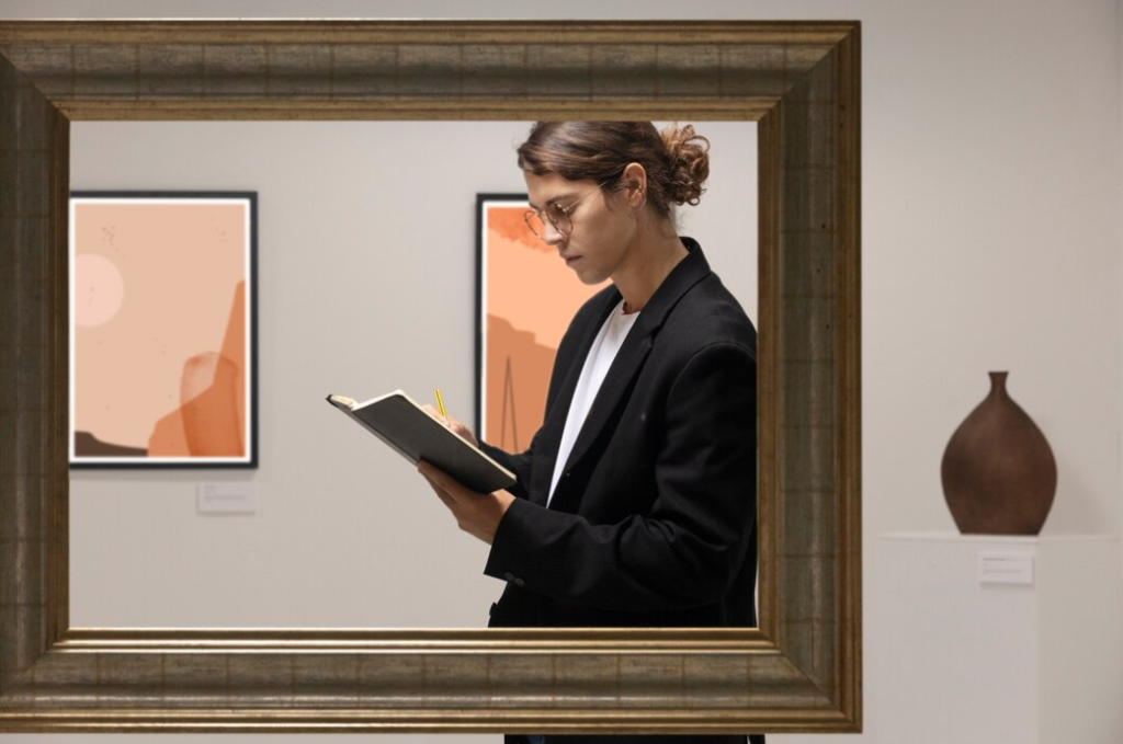 man with glasses standing in the frame with a book and pencil, the pictures on the wall behind him